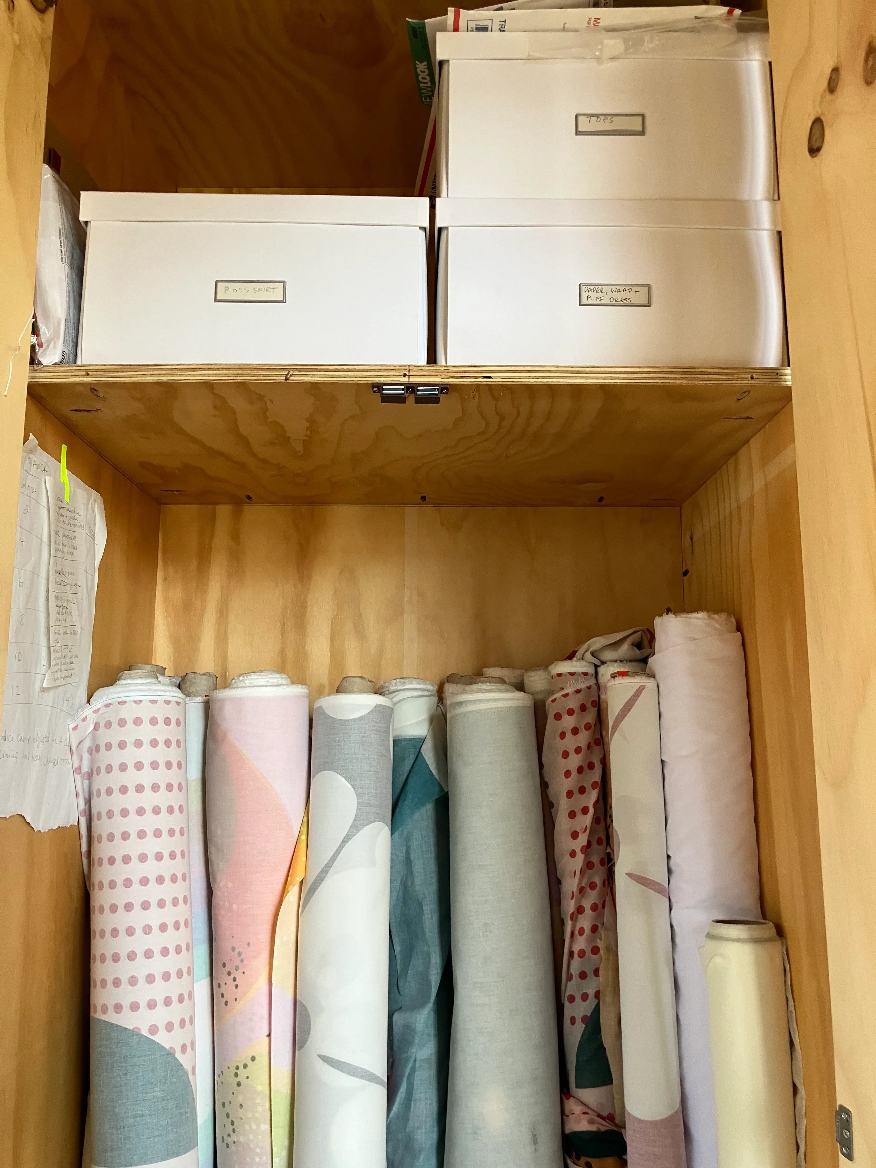 Colorful Hawaiian print fabric rolls stored in a wooden cabinet under labeled boxes.