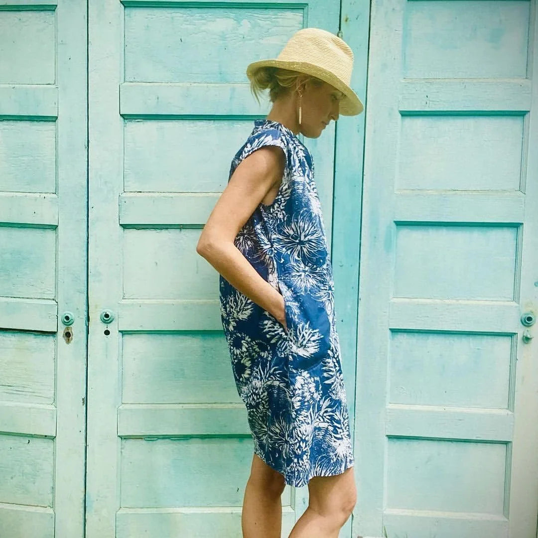 Woman in a blue floral Hawaiian mu'umu'u dress and straw hat stands by turquoise doors