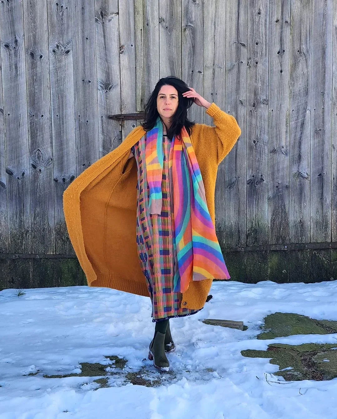 Woman in colorful mu'umu'u dress and mustard cardigan poses outside on snow by wood fence