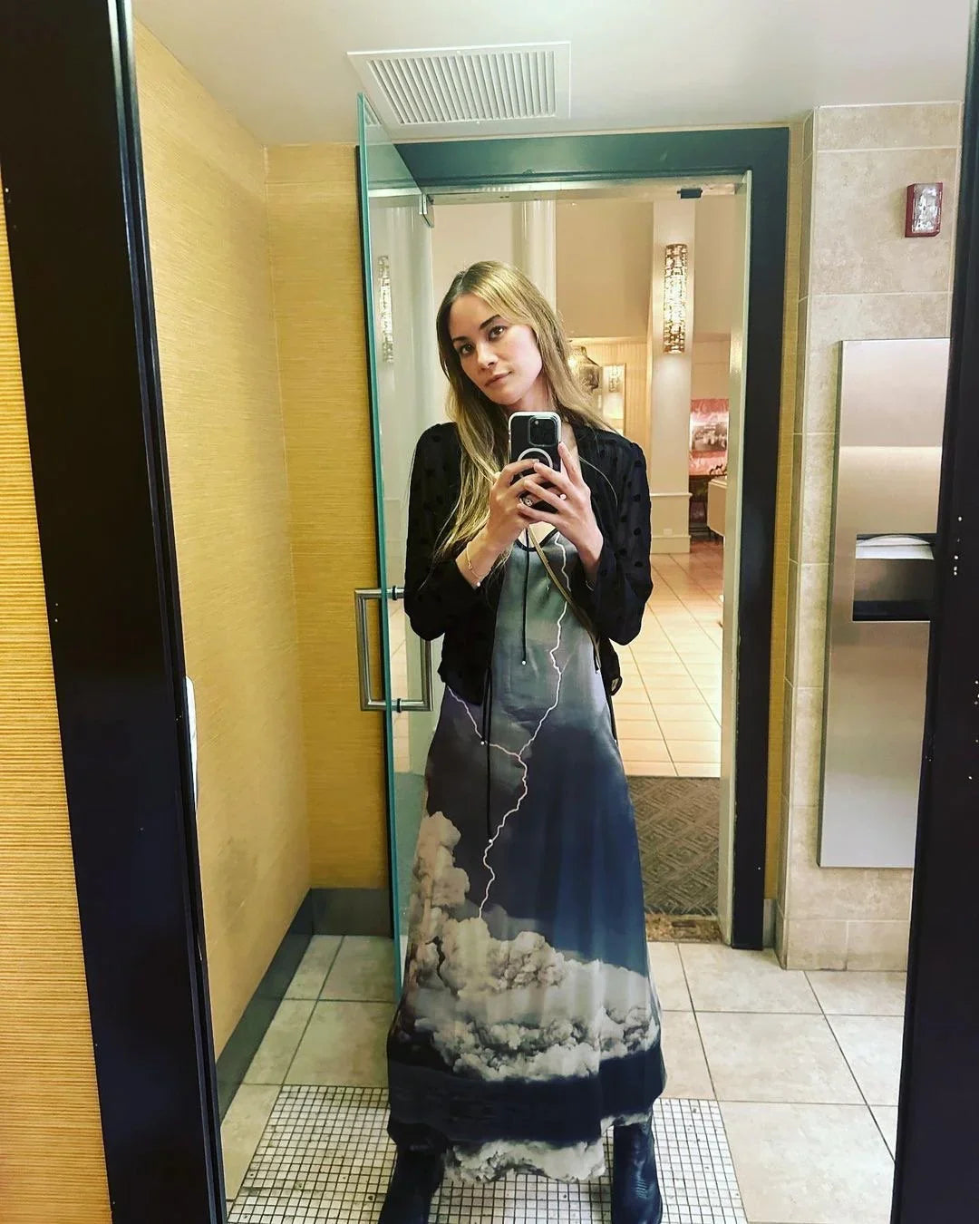 Woman wearing a long dress with a lightning and cloud print, taking a mirror selfie indoors.