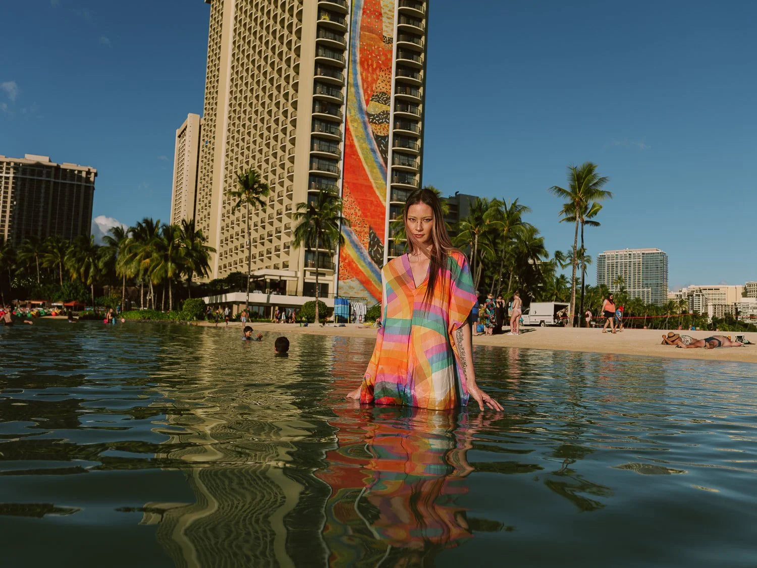 Woman in a colorful modern mu'umu'u dress standing in water with a Hawaiian beach backdrop.