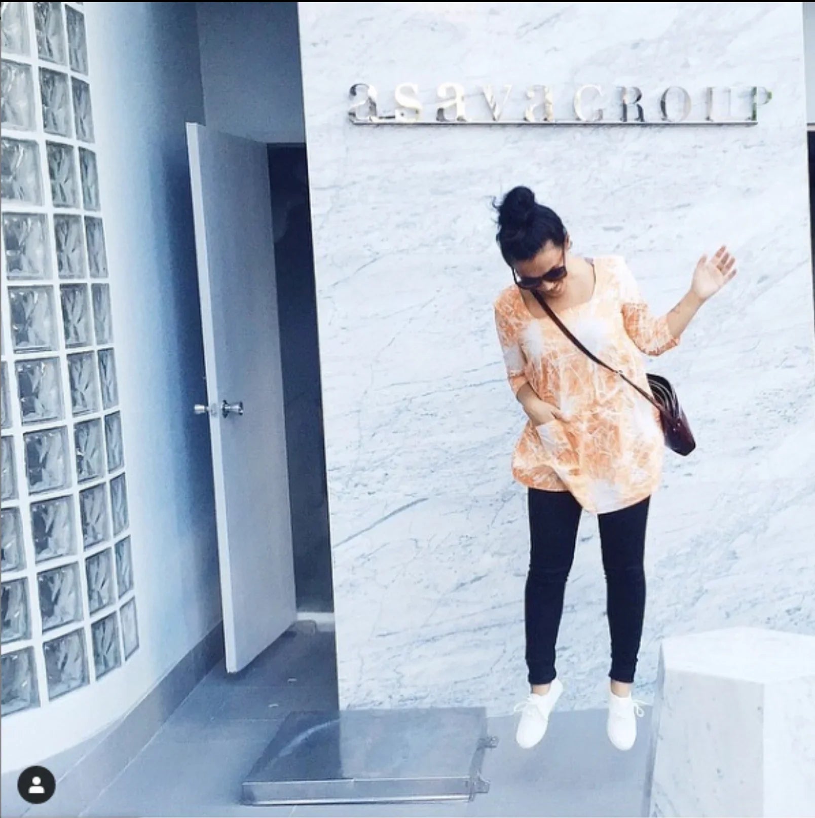 Woman in a modern mu'umu'u dress with orange Hawaiian print, black leggings, and white shoes posing against a marble wall.