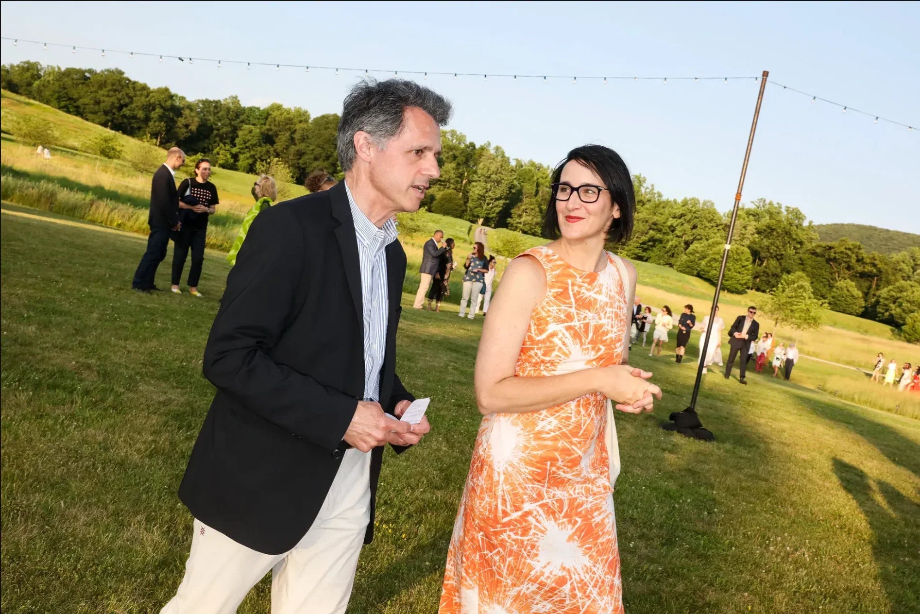 Woman in an orange floral muumuu dress outdoors at a summer gathering, grassy field background