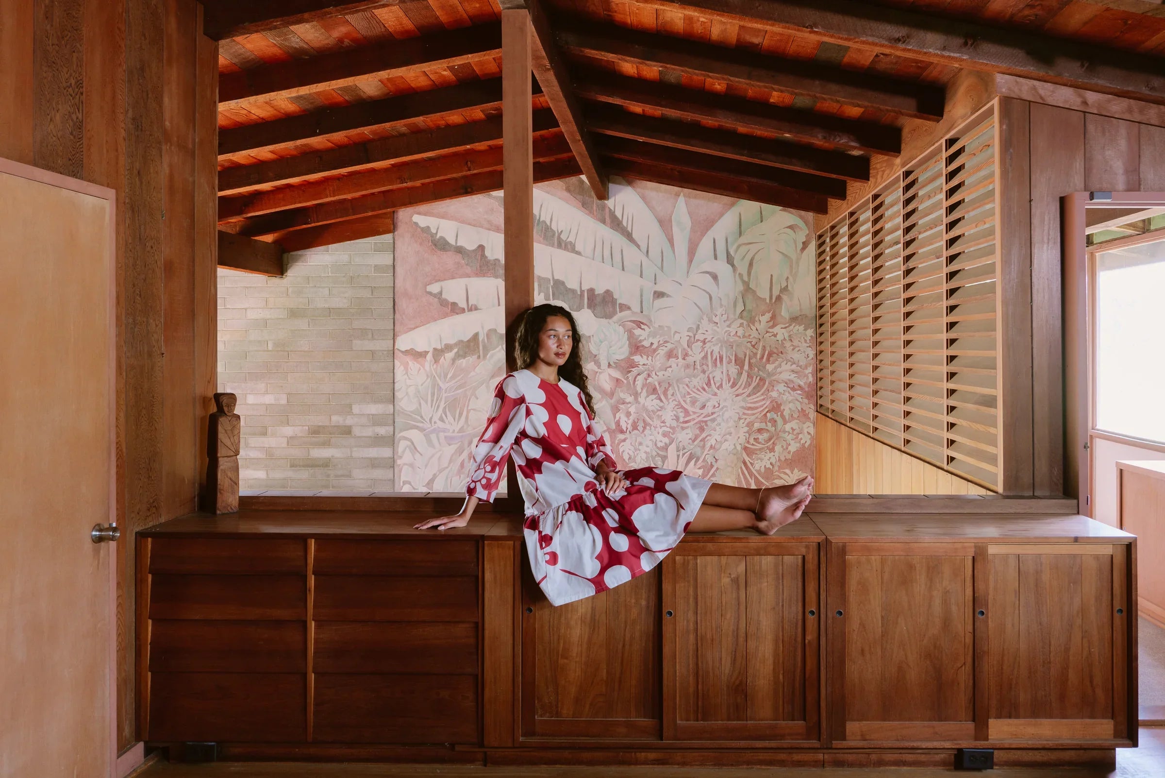 Woman in red and white muumuu dress poses indoors on wooden cabinet, tropical mural behind