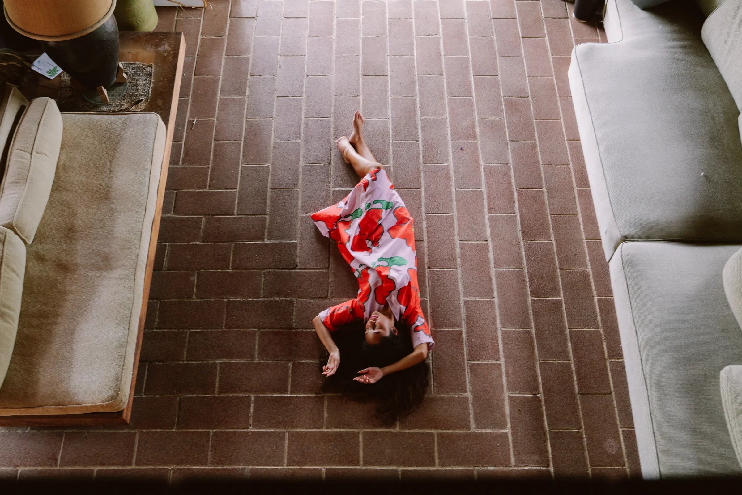 Woman in a modern floral mu'umu'u dress lying on a brick floor, surrounded by couches