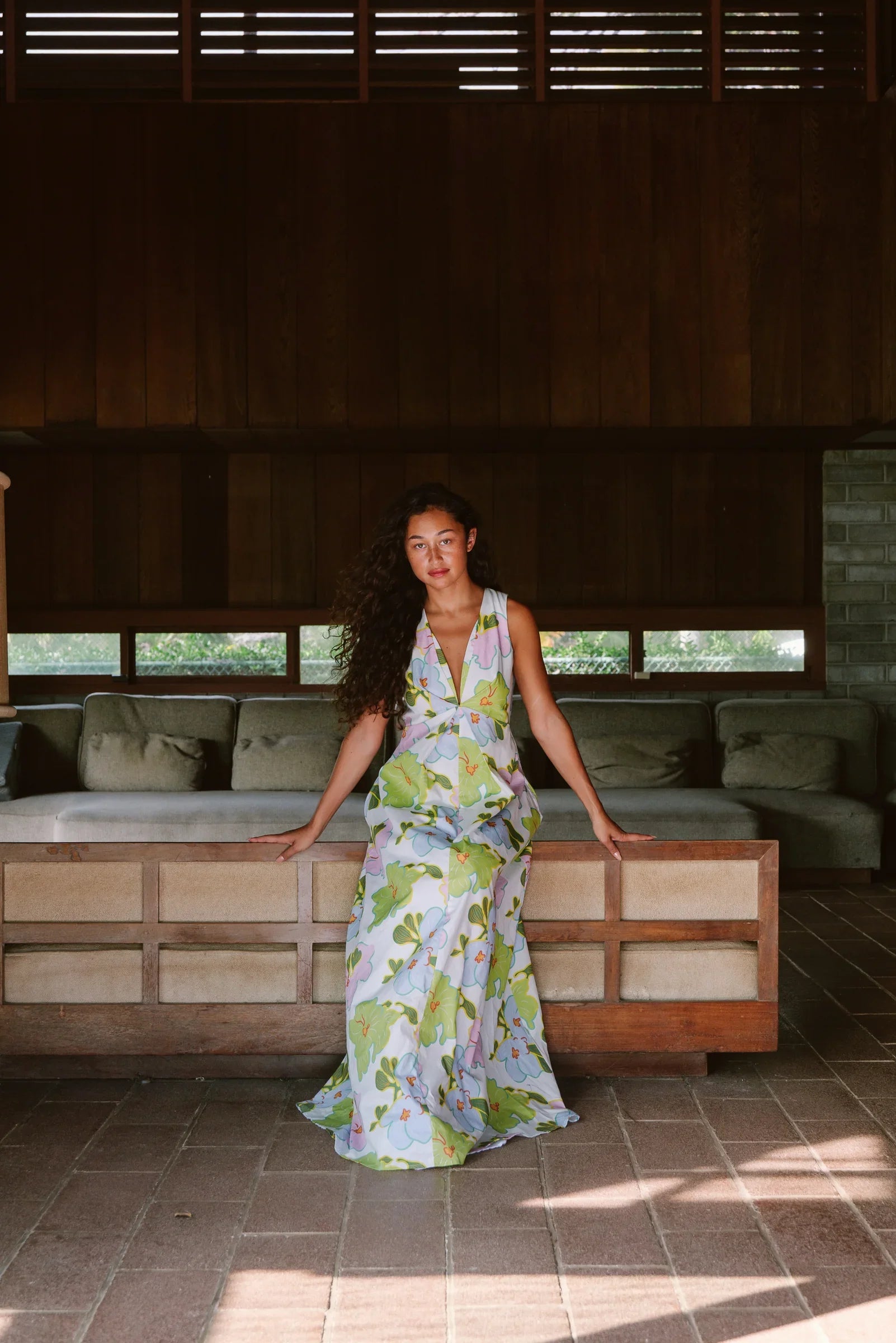Woman in a modern mu'umu'u dress with green and pastel floral Hawaiian print sitting indoors