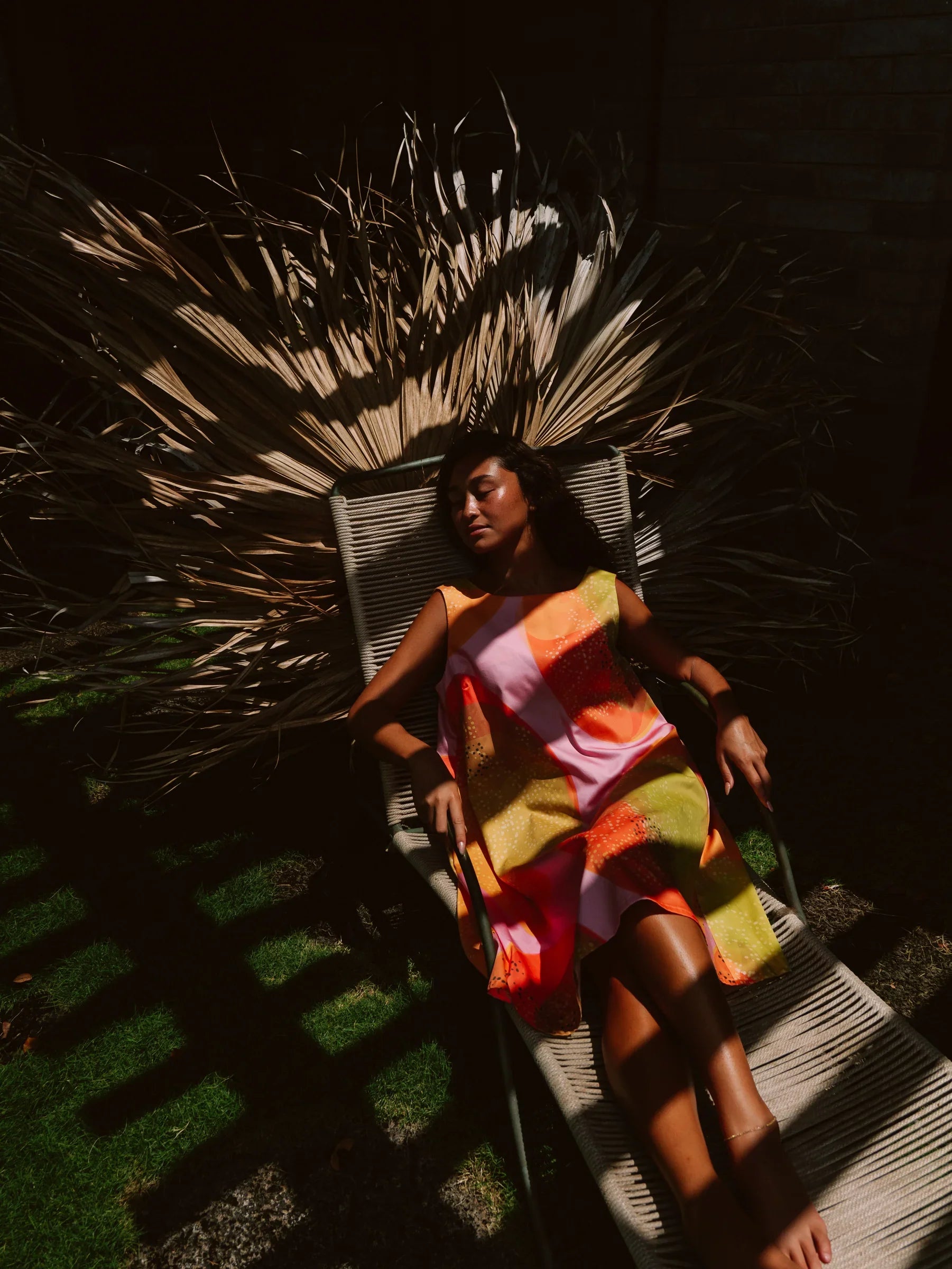 Woman in a colorful modern mu'umu'u dress relaxing outdoors on a lounge chair with palm leaves