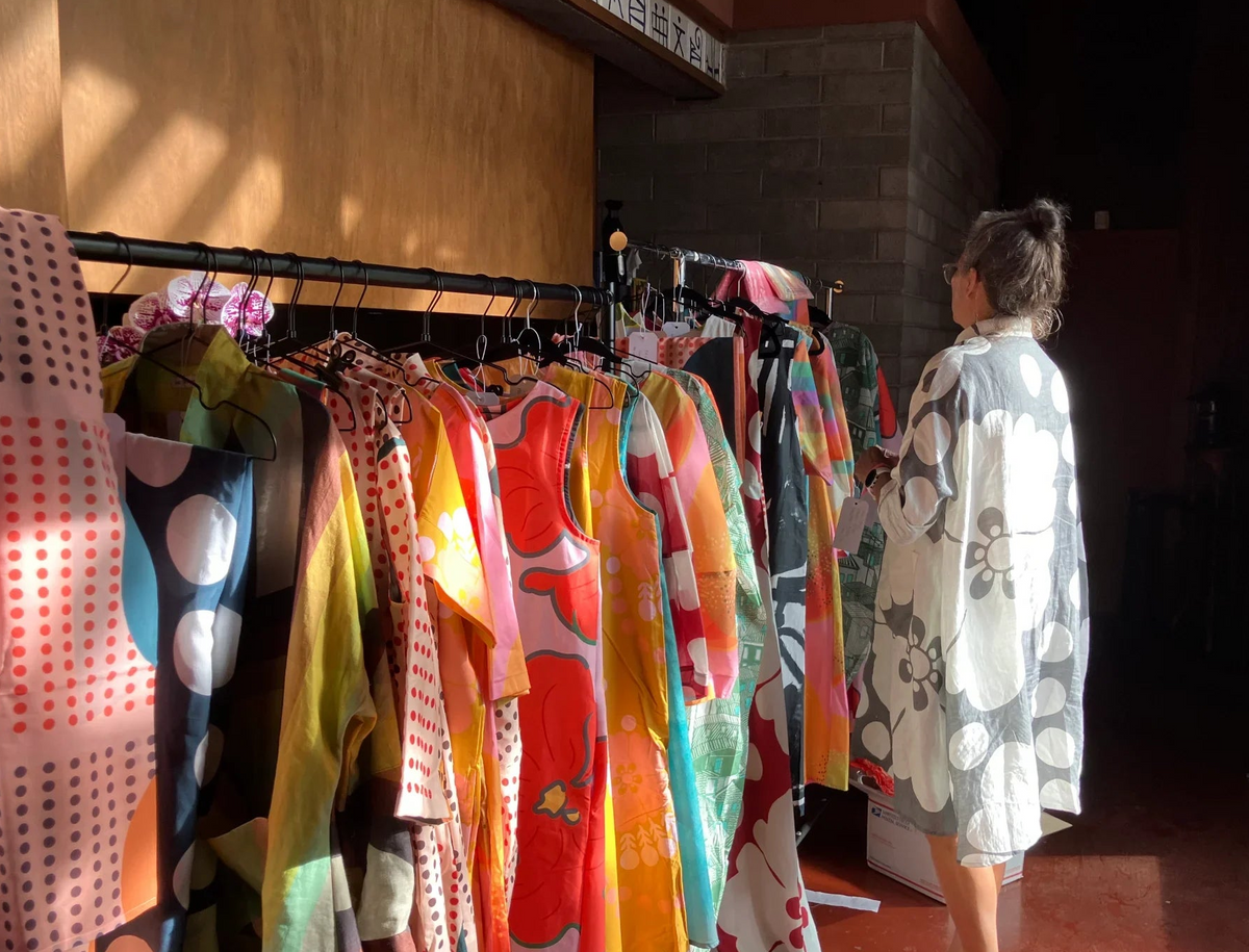 A woman wearing a floral pattern dress stands beside a clothing rack filled with colorful patterned garments in a shop interior.