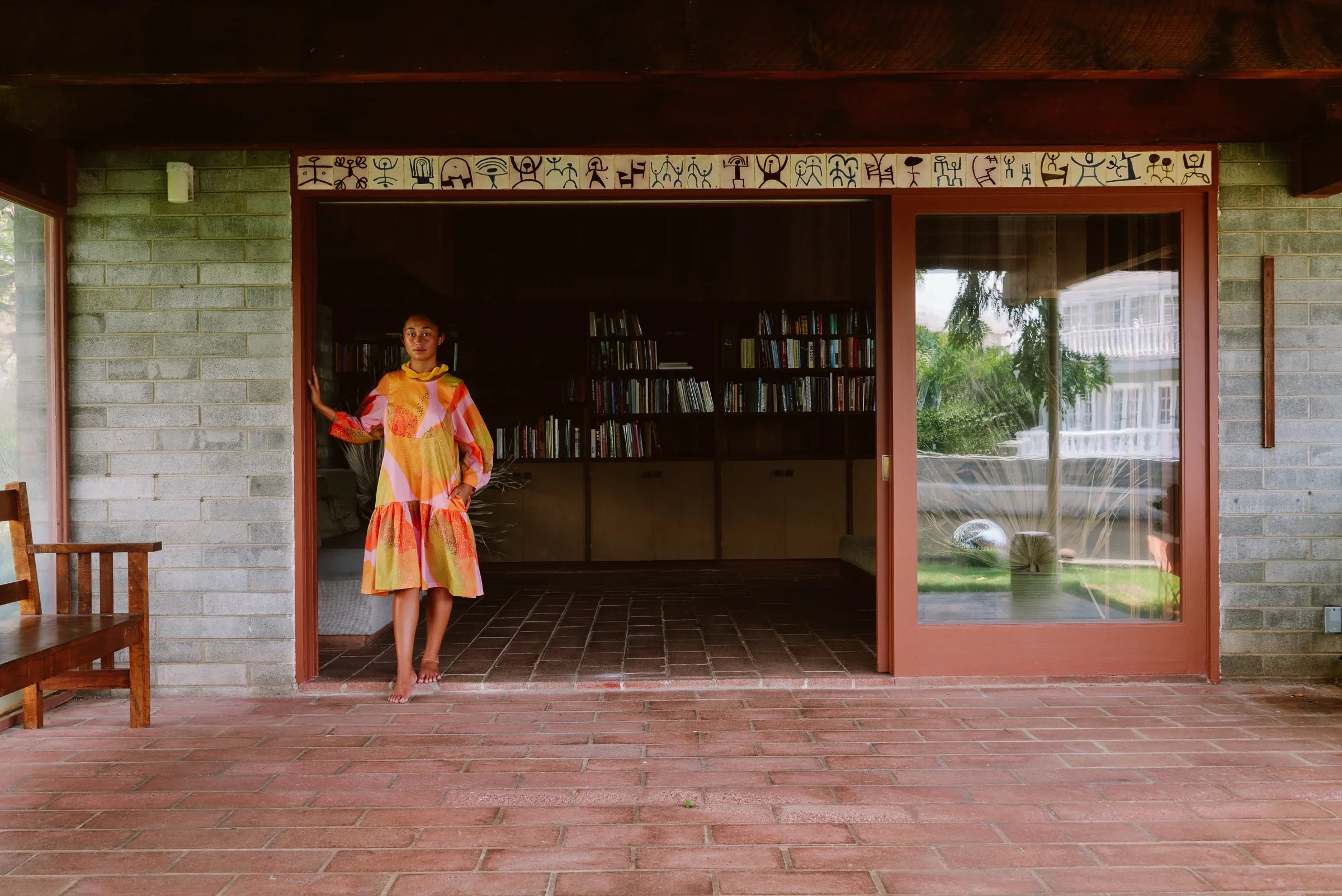 Woman in a colorful modern muumuu dress stands barefoot in a doorway with bookshelves inside.