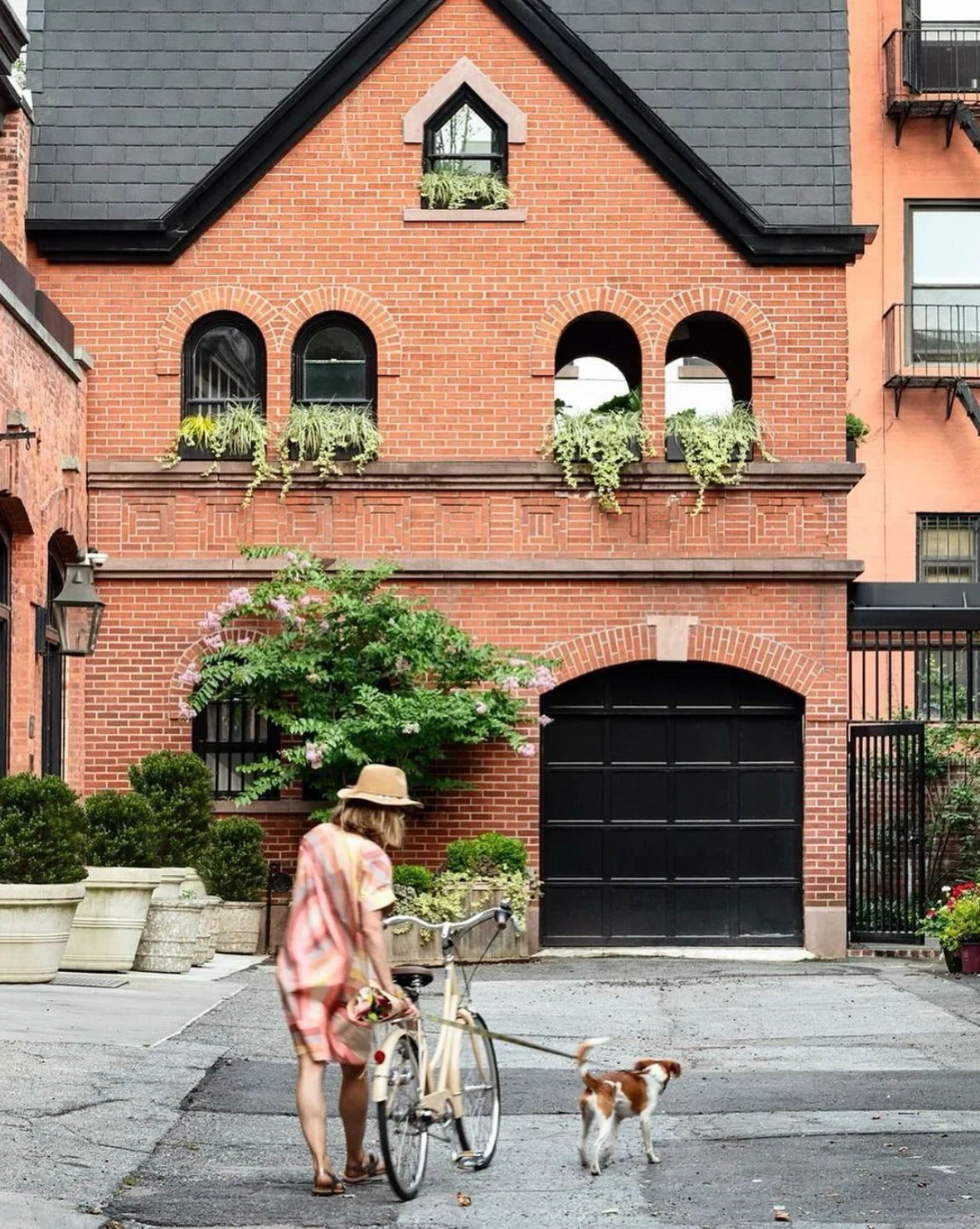 Woman in a modern mu'umu'u dress walking a dog with a bicycle in front of a brick house
