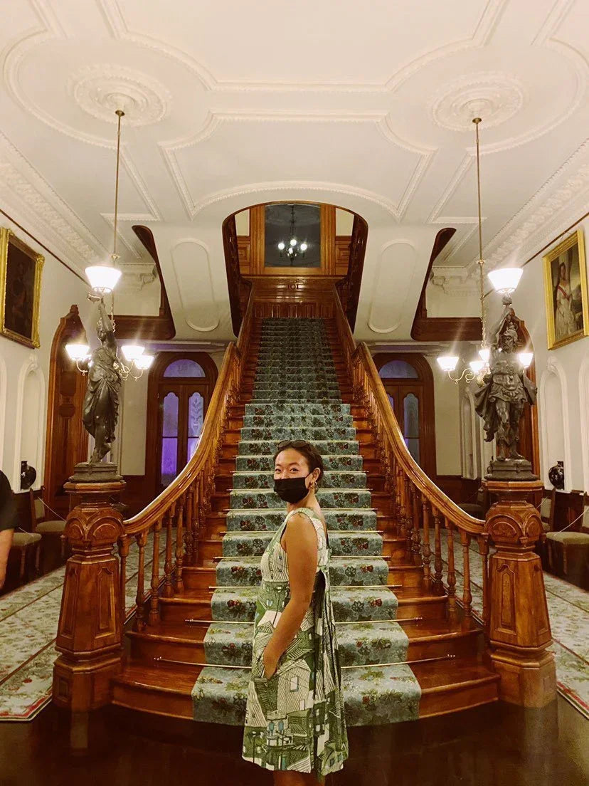 Woman in a modern green Hawaiian mu'umu'u dress stands masked by ornate grand staircase