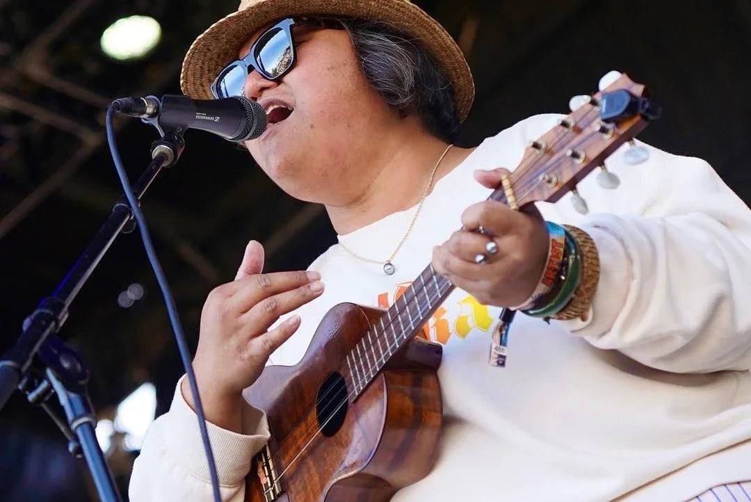 Person singing into microphone and playing ukulele on stage, wearing hat and sunglasses