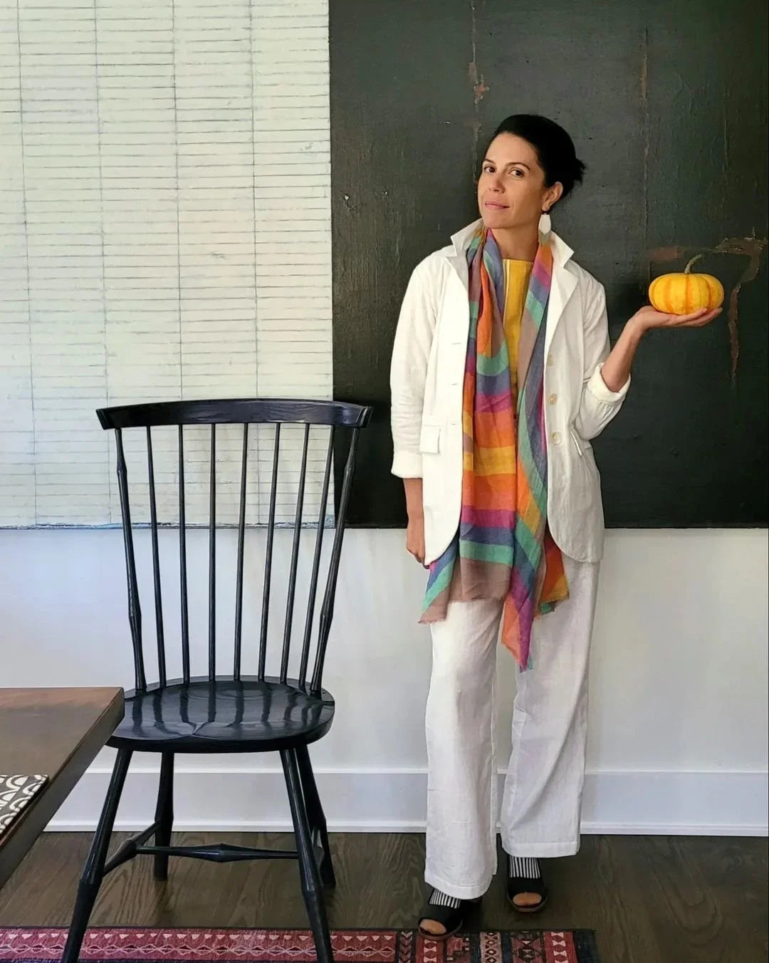 Woman in white outfit with colorful striped scarf holding a small pumpkin, standing indoors