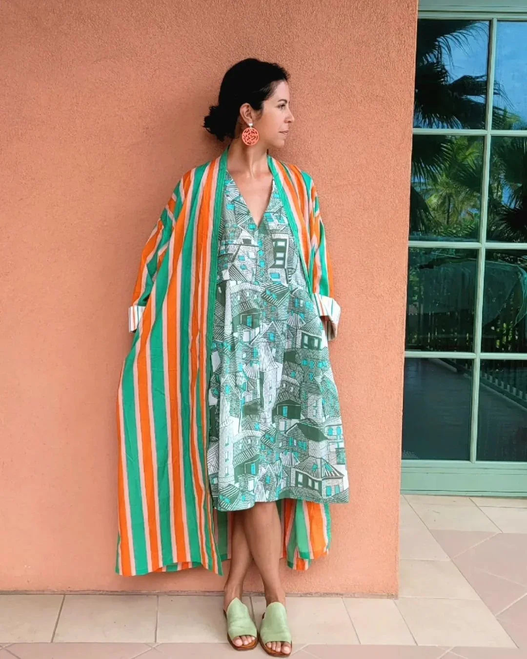 Woman in a green and orange modern mu'umu'u dress with Hawaiian print standing by a peach wall