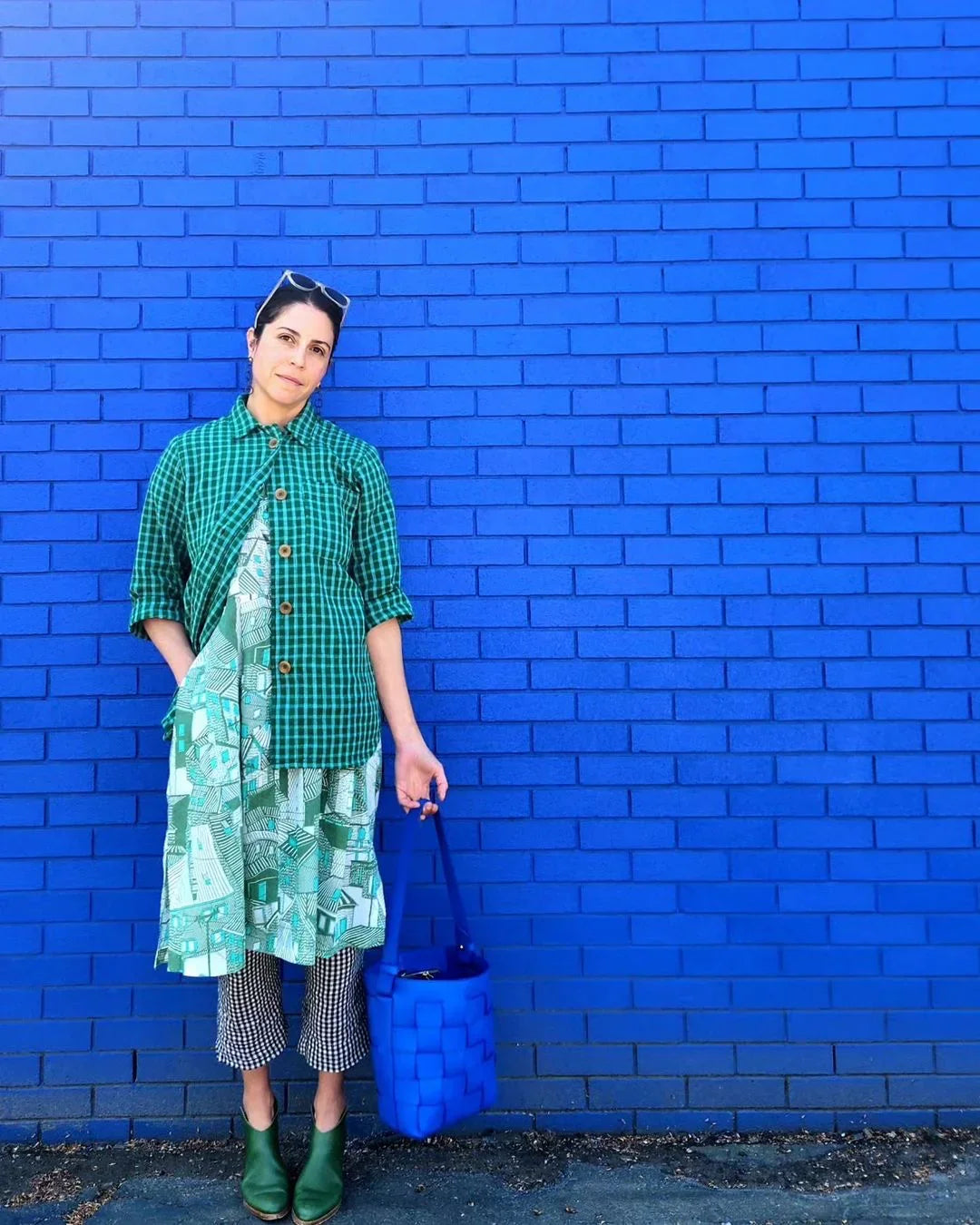 Woman in layered modern mu'umu'u dress, green plaid and Hawaiian prints, stands by blue brick wall
