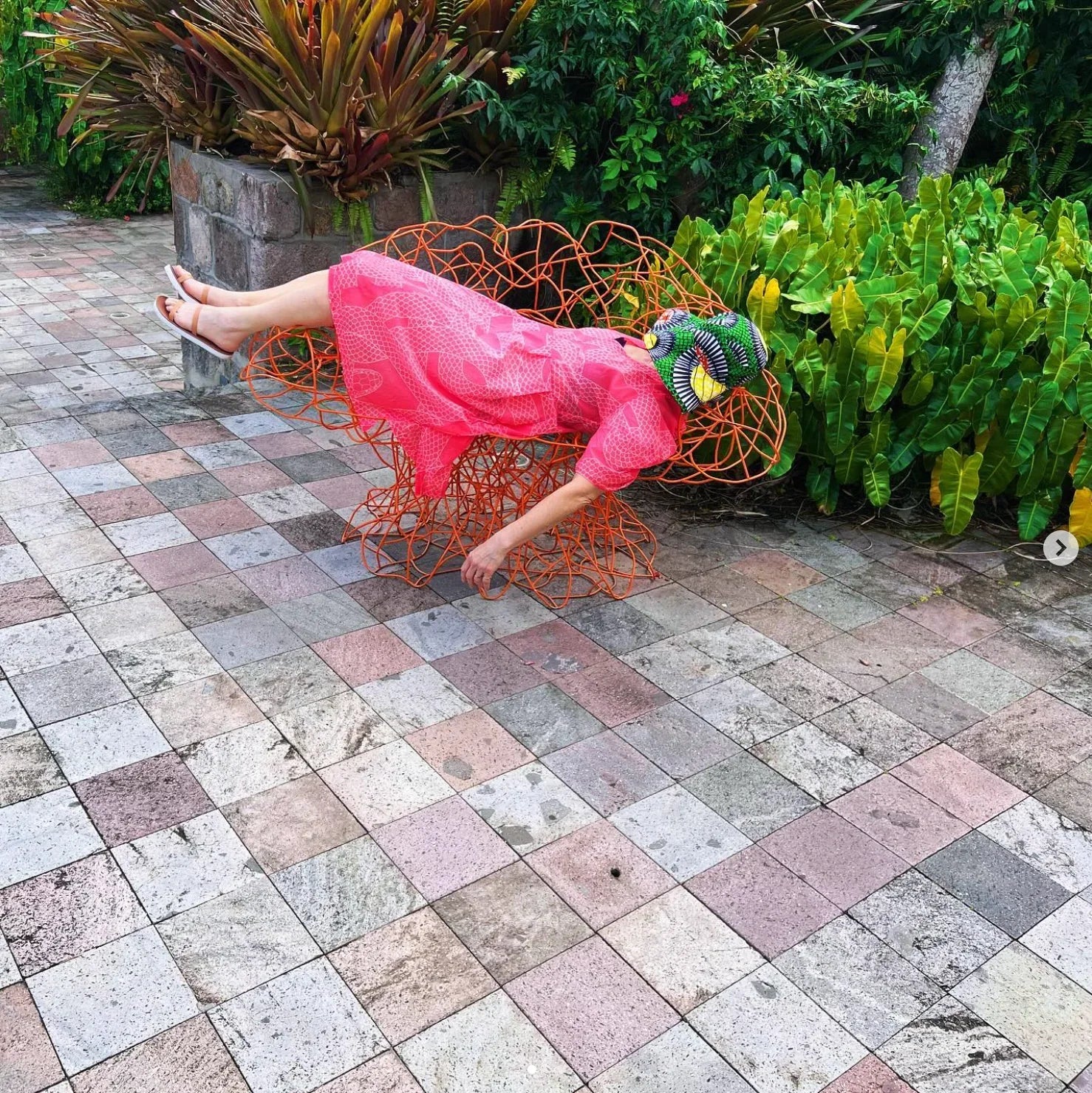Woman in a pink modern muumuu dress lounging on orange wire chair in tropical outdoor garden