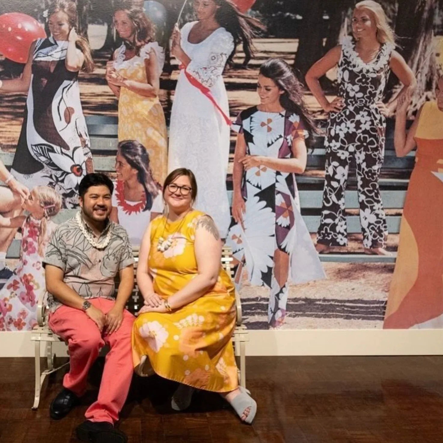 Two people in modern Hawaiian print outfits sitting before a mural of vintage mu'umu'u dresses.
