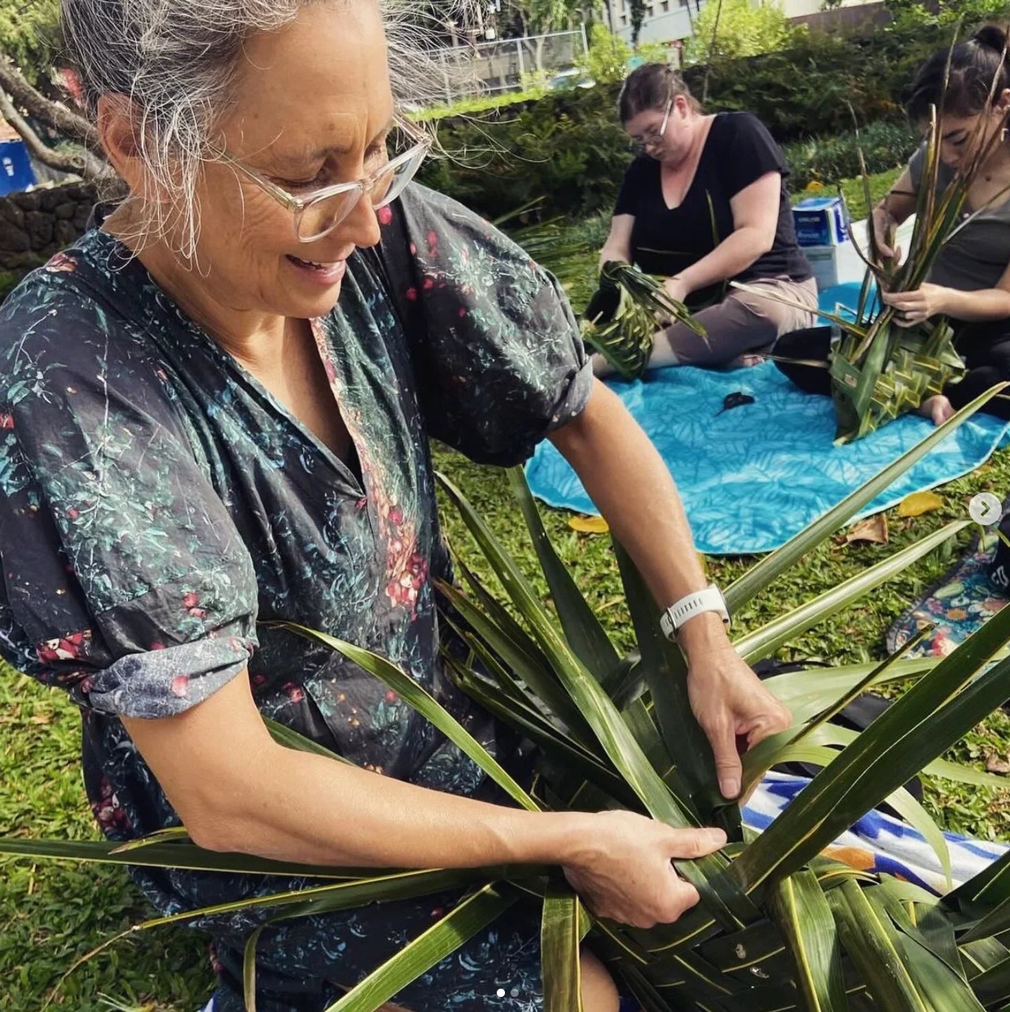 Woman in a modern mu'umu'u dress weaving palm fronds outdoors with others on grass.