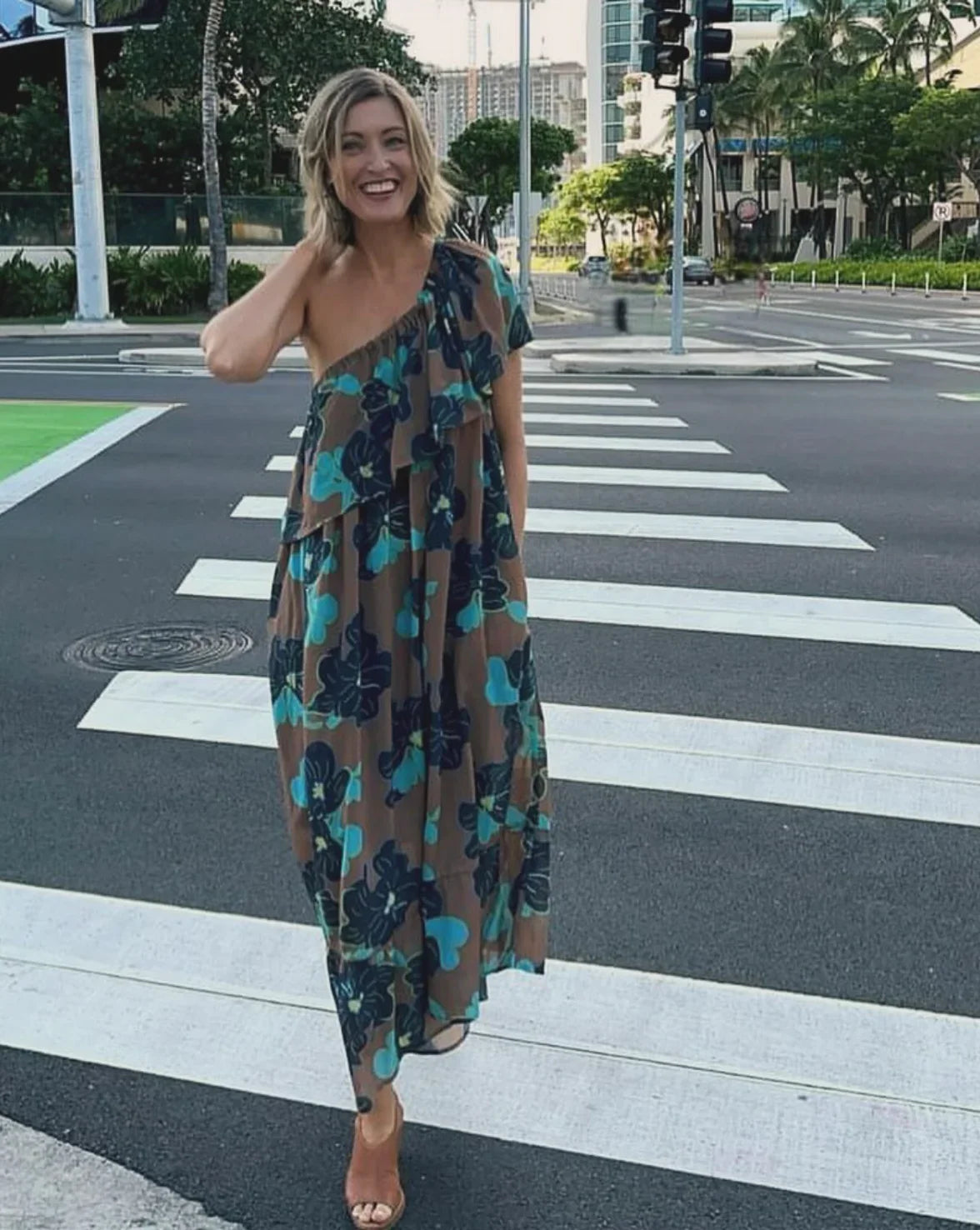 Woman wearing a modern one-shoulder mu'umu'u dress with blue Hawaiian floral print, standing on a city crosswalk.