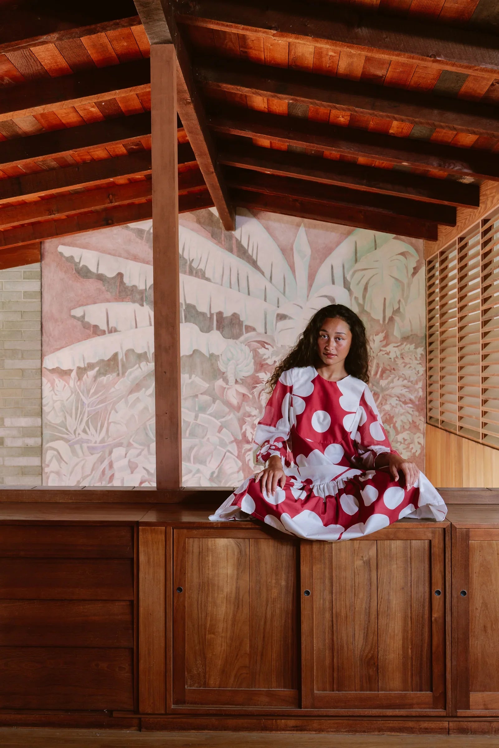 Woman in a red and white polka dot modern muumuu dress sitting on wooden cabinet, tropical mural background