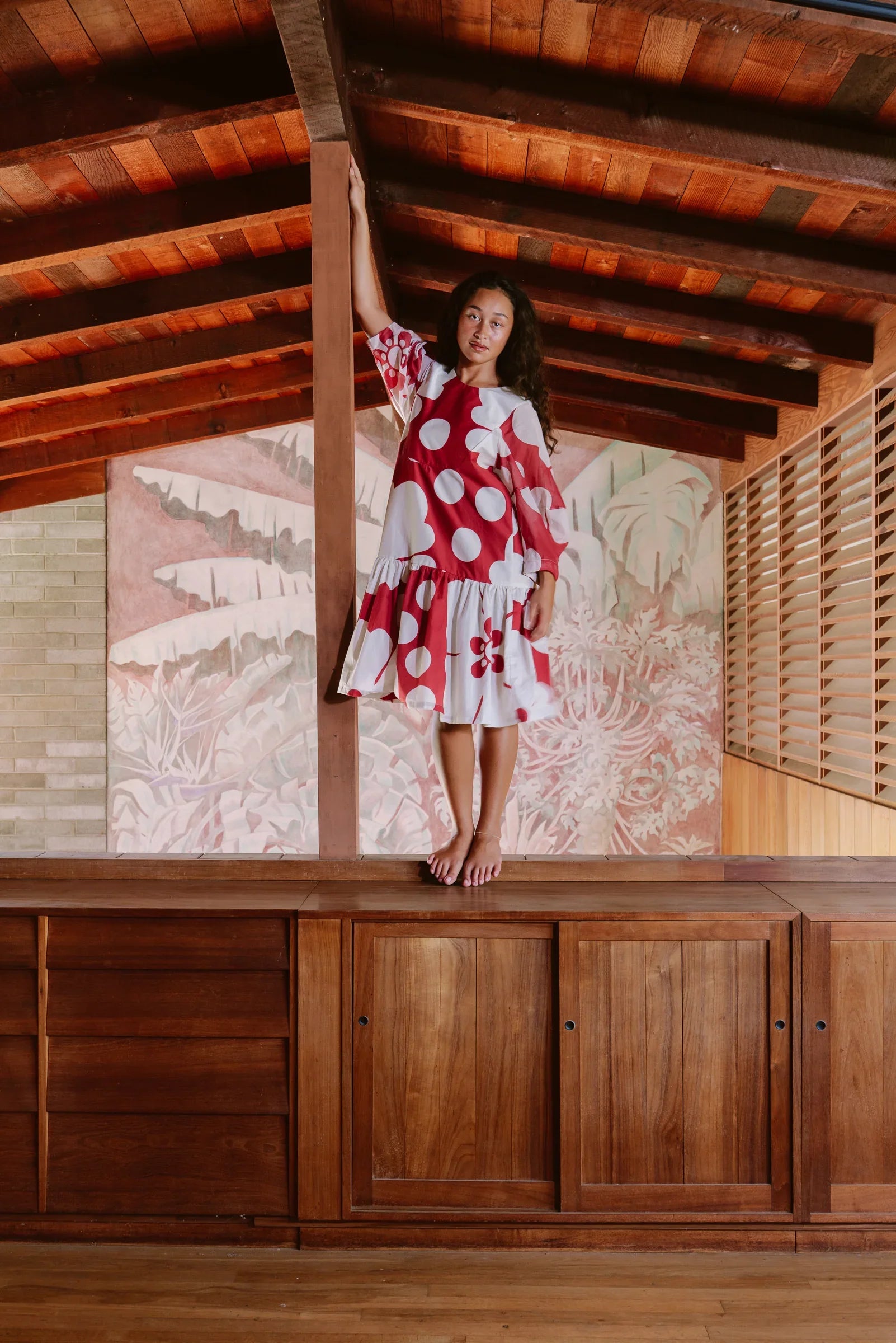 Woman in a red and white polka dot modern mu'umu'u dress stands barefoot indoors with wood accents