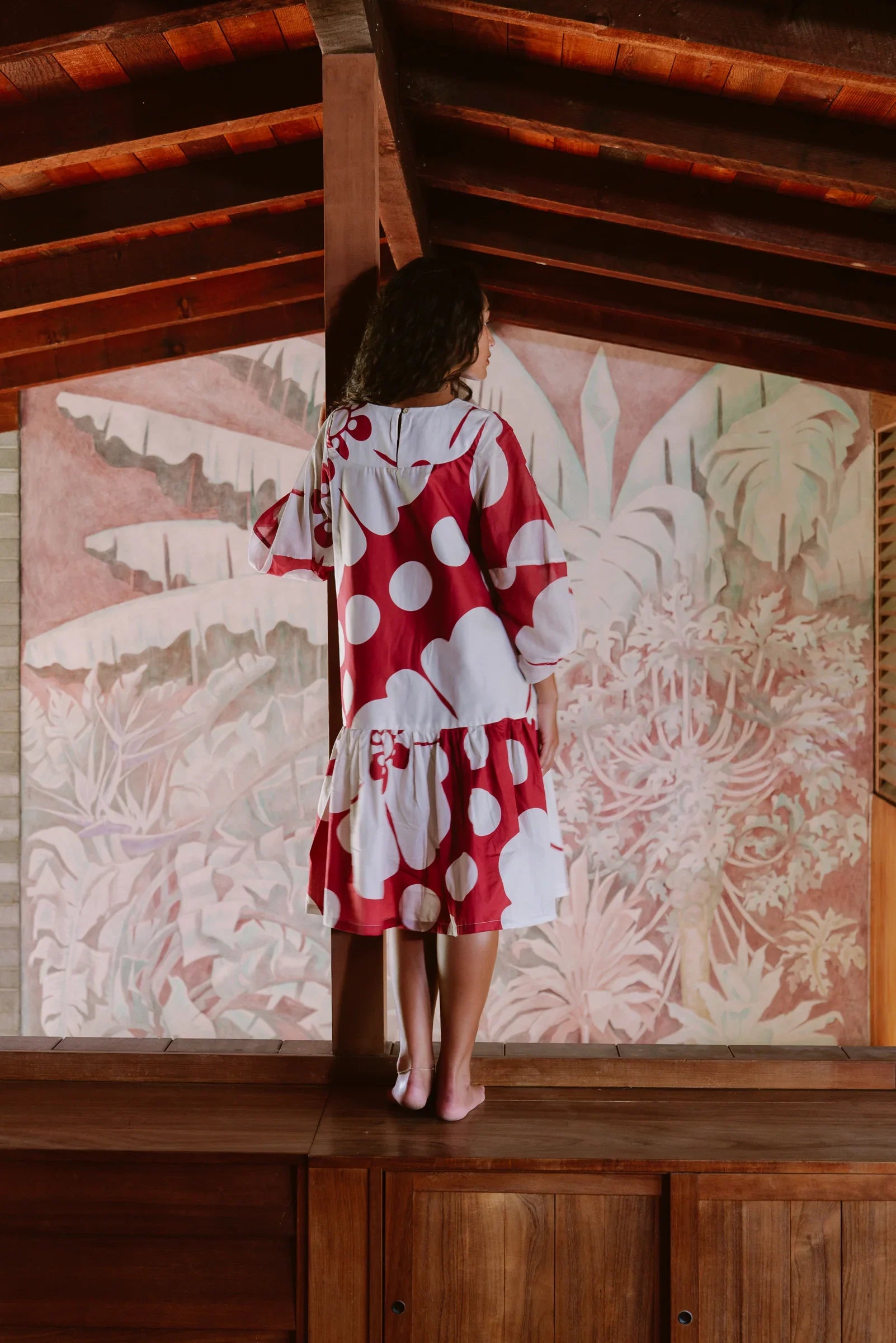 Woman in red and white polka dot mu'umu'u dress standing indoors by tropical mural