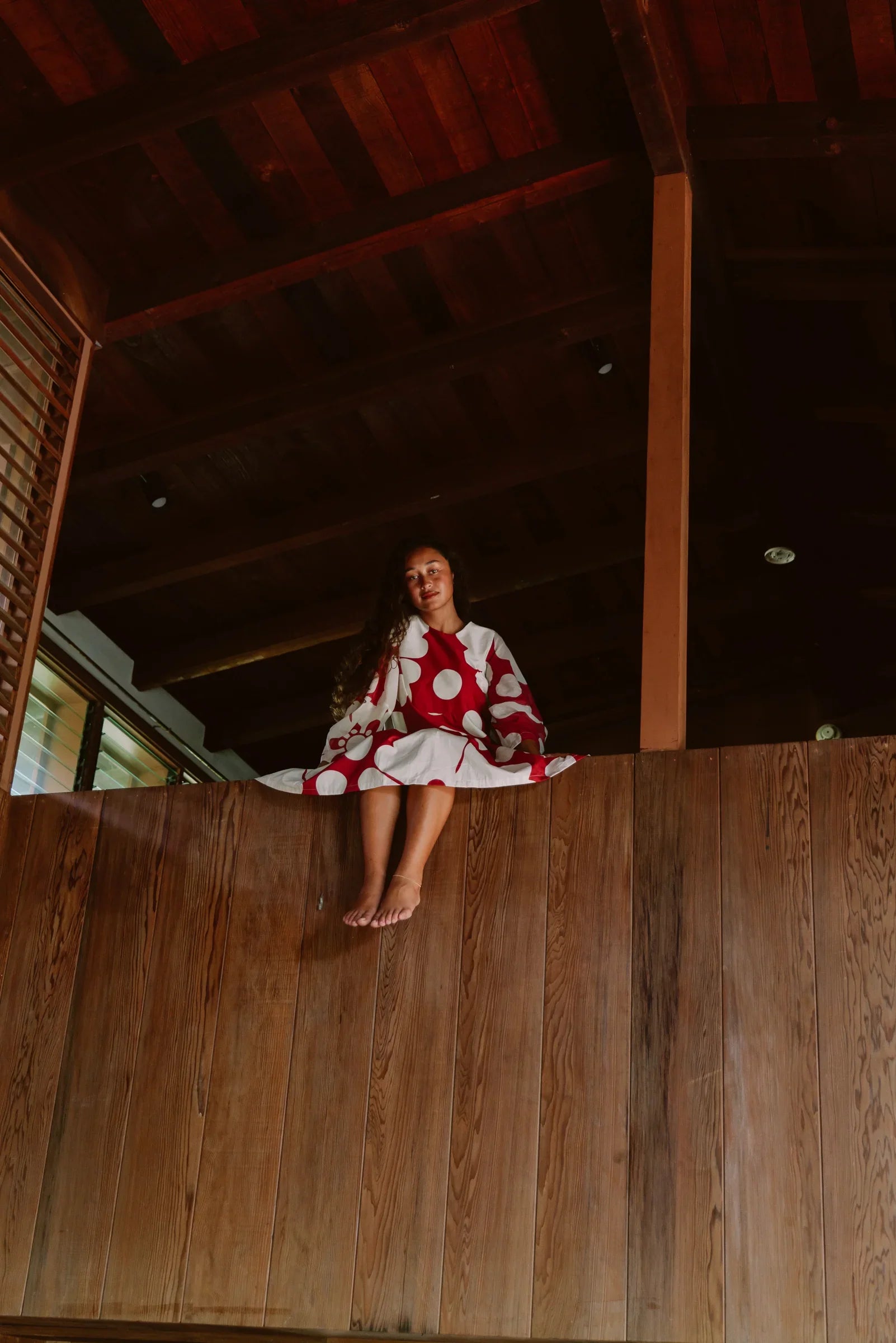 Woman in a red and white modern mu'umu'u dress sitting on a wooden ledge indoors