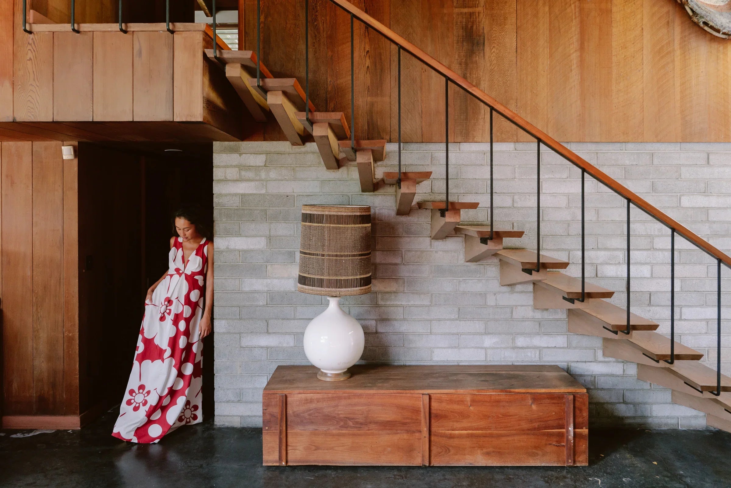 Woman in a red and white floral modern mu'umu'u dress stands by stairs in a mid-century room