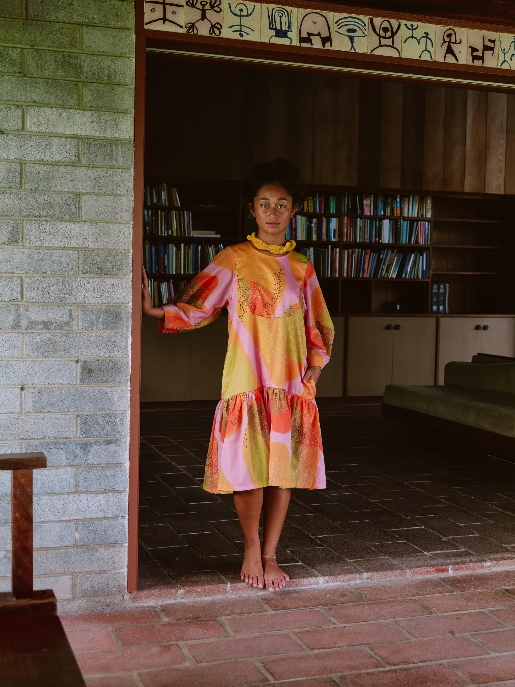 Woman in a colorful modern mu'umu'u dress with abstract Hawaiian prints, standing barefoot indoors