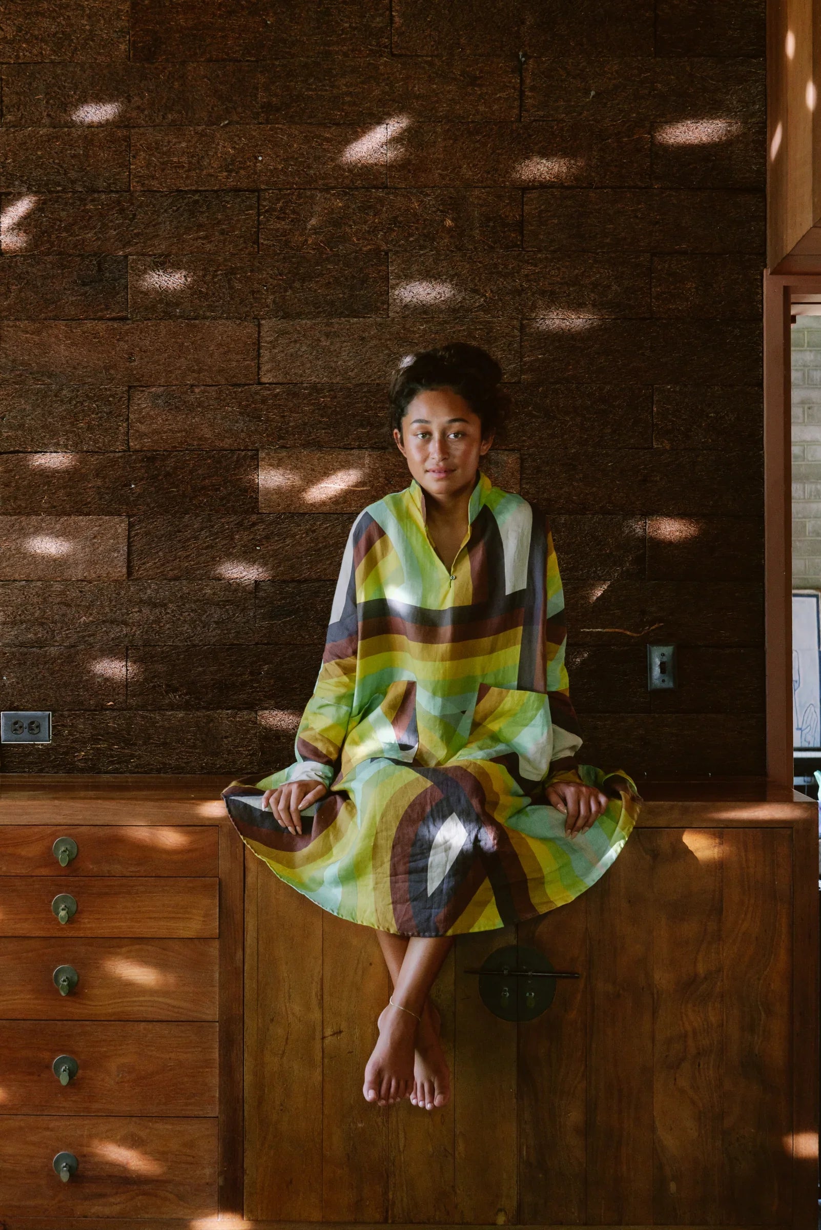 Woman in a colorful modern mu'umu'u dress sitting indoors against a textured brown wall