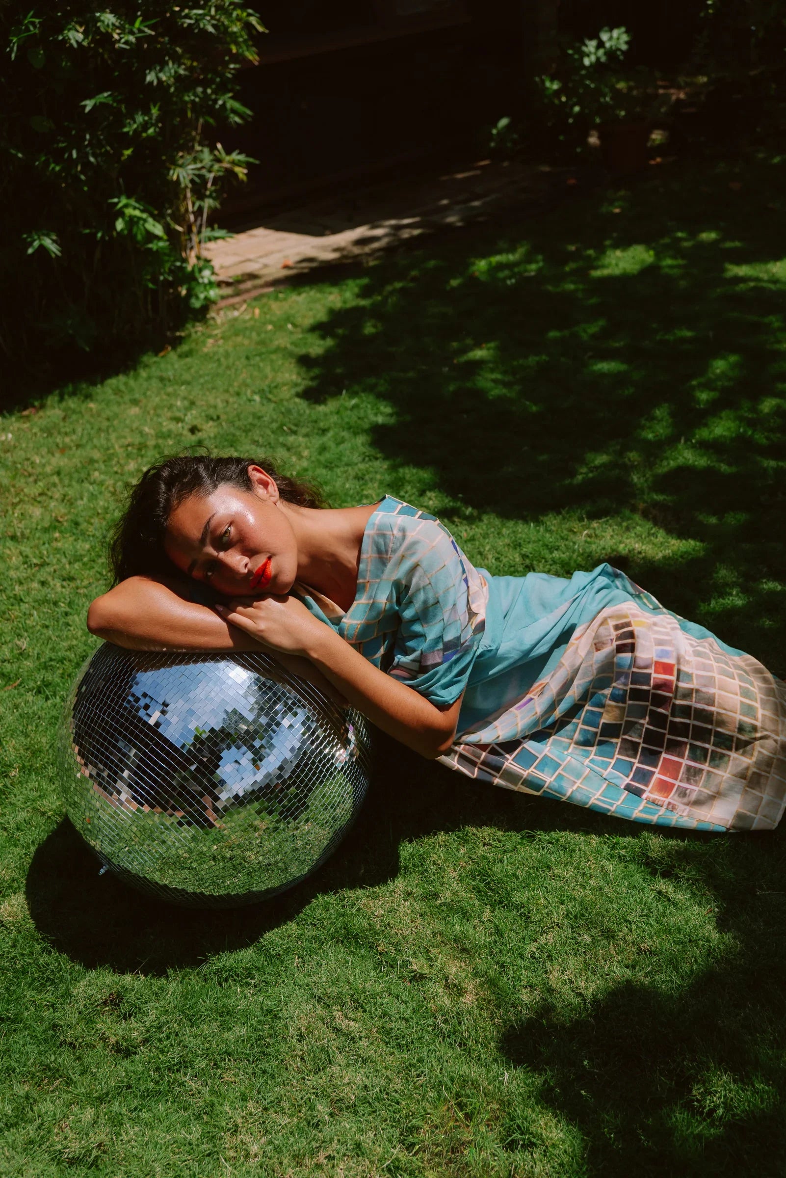 Woman in modern mu'umu'u dress with grid pattern lying on grass beside a disco ball
