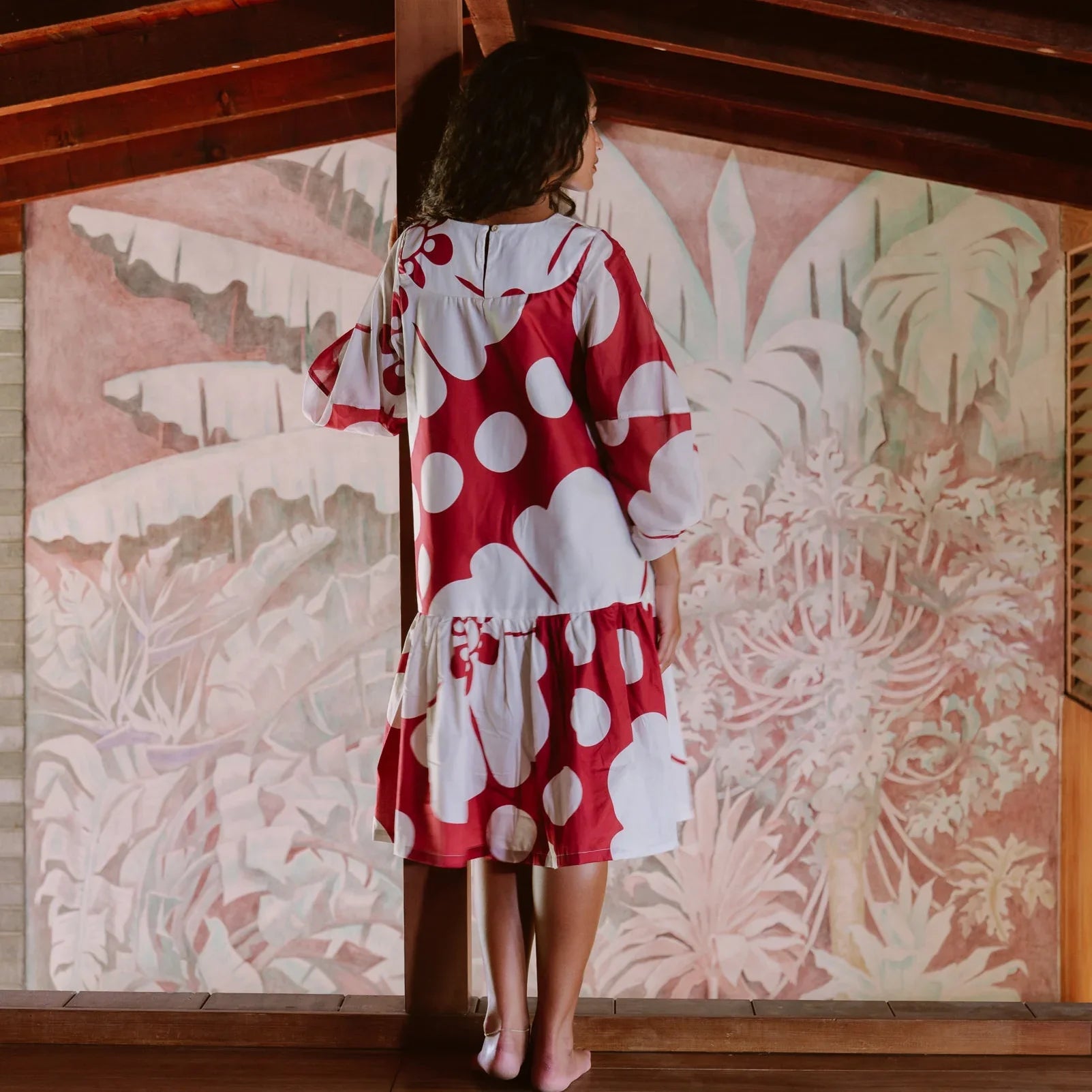 Woman in a red and white polka dot muumuu dress standing indoors by tropical mural