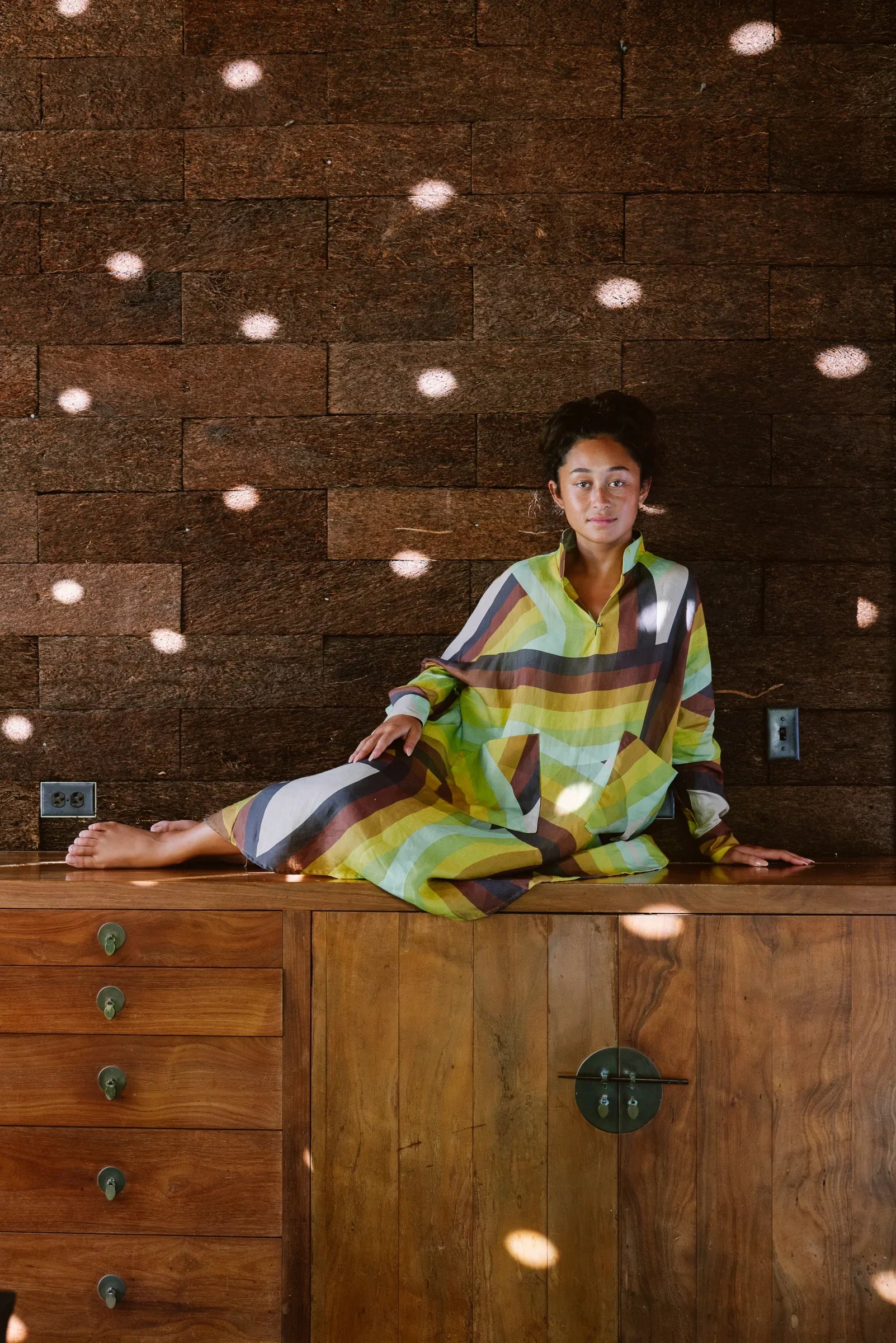 Woman in a modern muumuu dress with bold geometric stripes, seated indoors against a wooden wall.