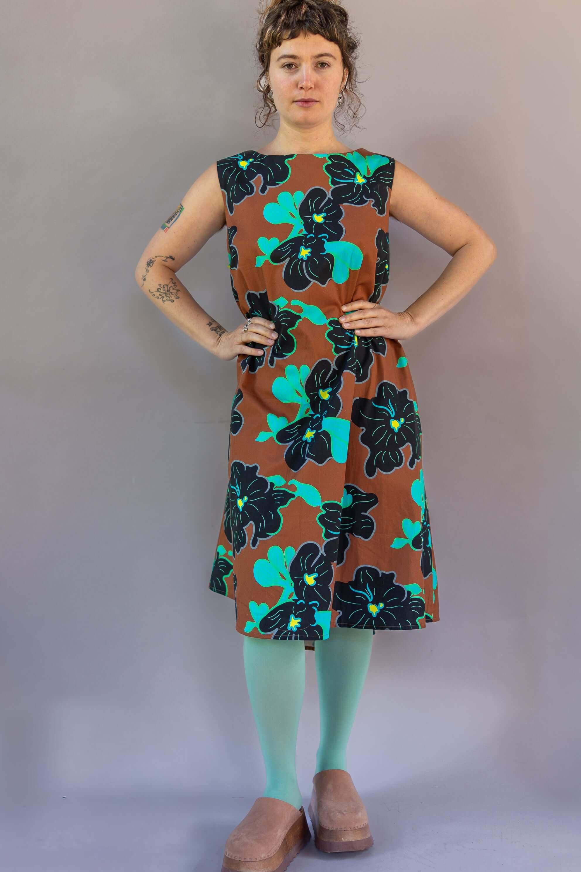 Woman wearing a brown sleeveless dress with bold teal and black floral print, teal tights, and tan shoes