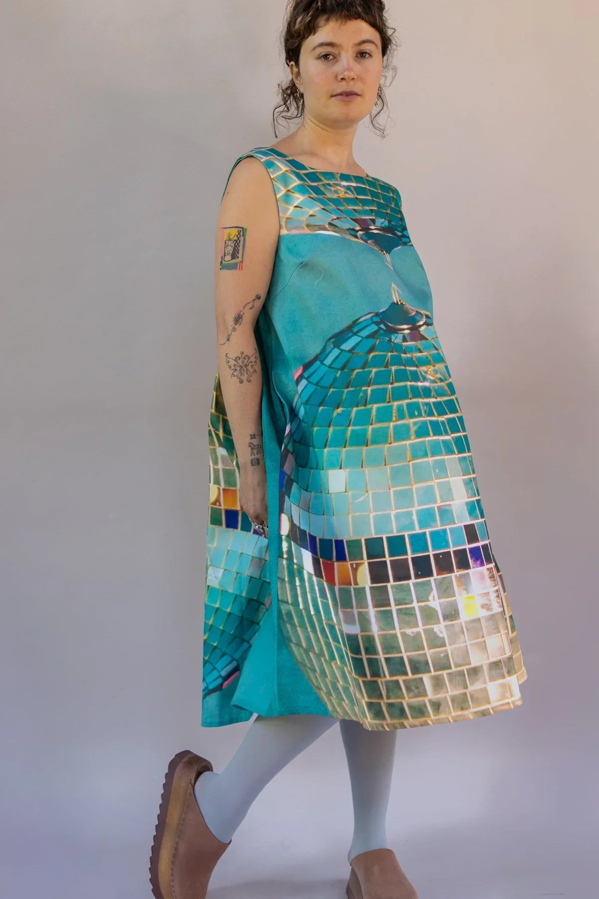 Woman models teal modern muumuu dress with bold disco ball print, casual shoes and tattoos.